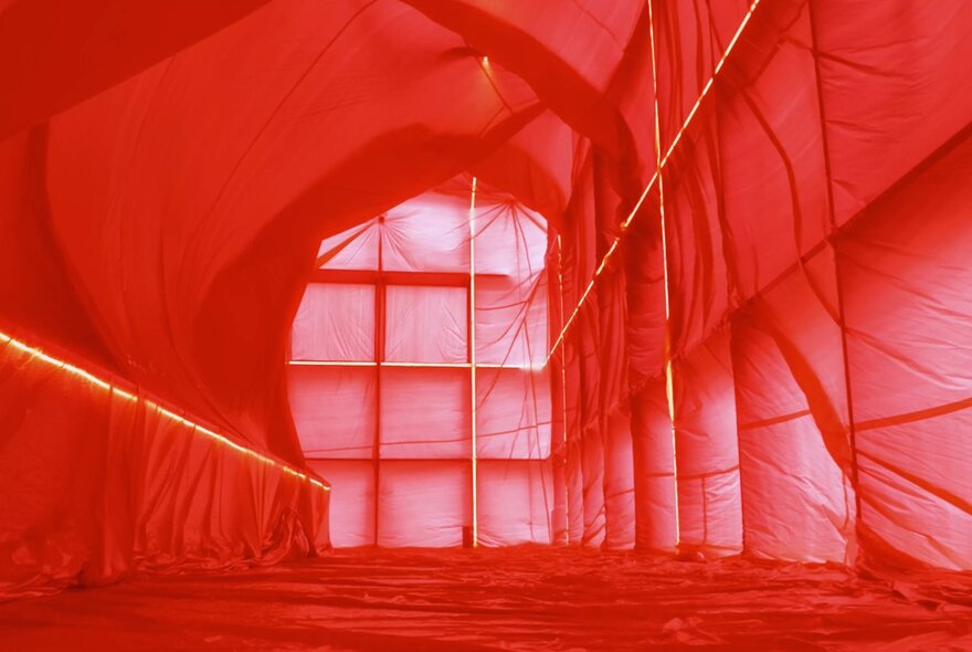 A large interior space, with sheer fabric draped along the walls, floor and ceiling, the space bathed in a red light.