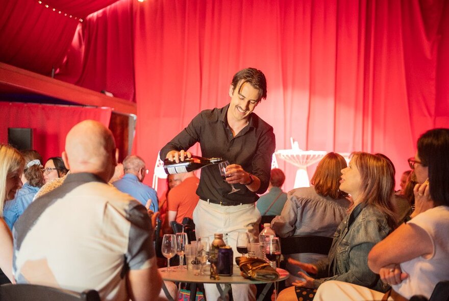 A man walking among a seated audience at a comedy festival gig and pouring wine from a bottle into a glass.