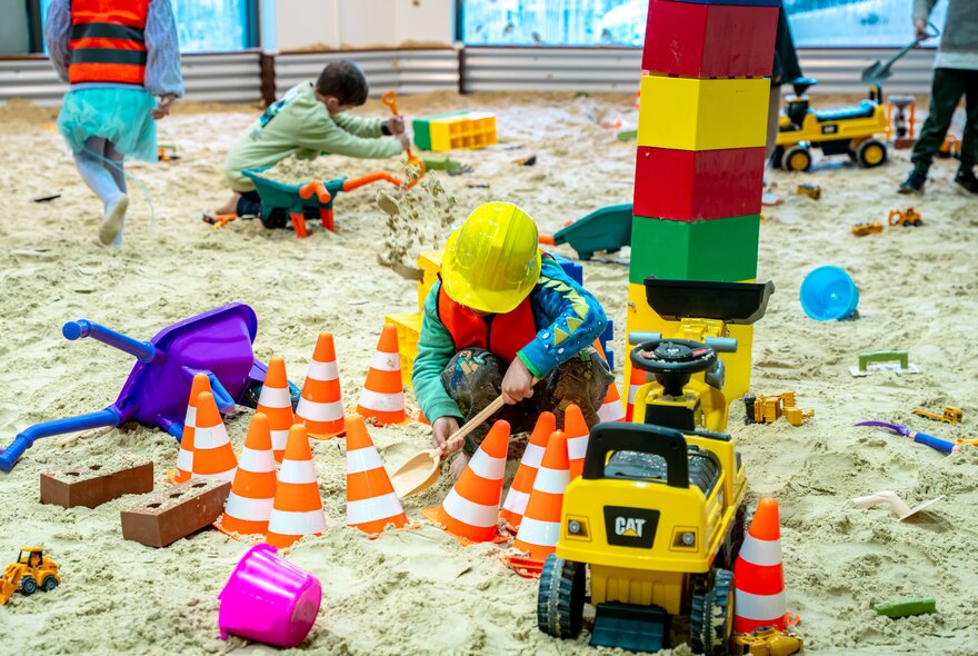 Children in a large indoor sandpit filled with diggers, mini-witch's hats and large building blocks.