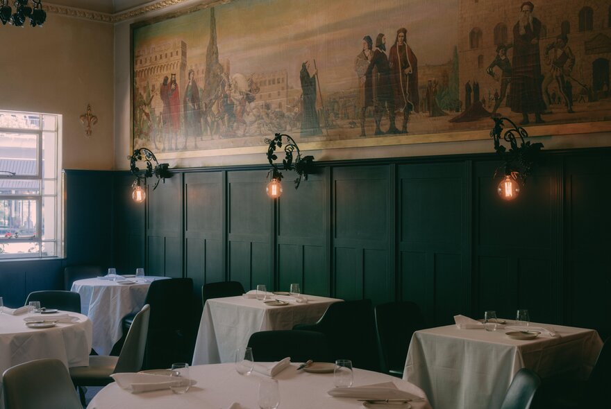 An elegant, dimly lit restaurant dining room featuring dark wood panelled walls beneath a long, historic mural depicting historical figures; three vintage wall sconces with glowing light bulbs illuminate white tablecloth-covered dining tables neatly arranged across the room.