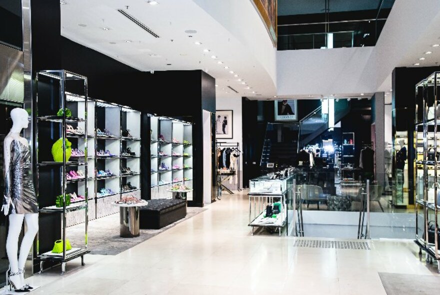 Interior of Harrolds store, a large open space, white walls, white ceiling and tiled floor, with racks of glass shelves to the left displaying leather bags.