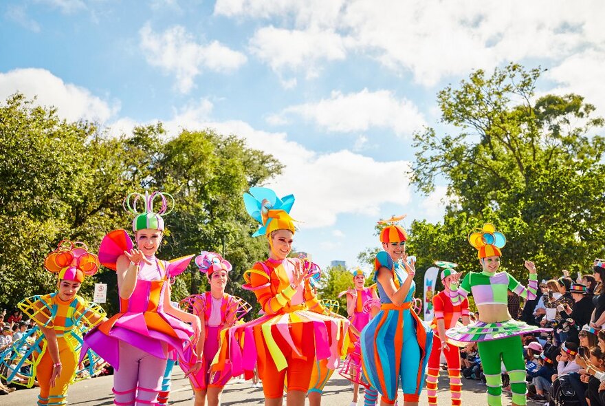 Troupe of people in brightly coloured costumes taking part in a street parade, with onlookers watching. 