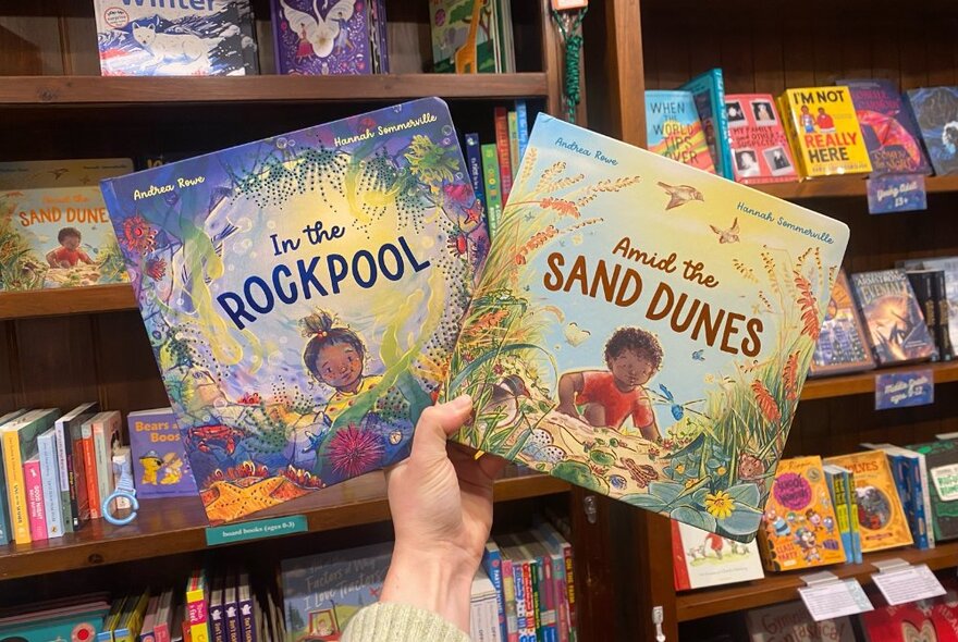 A hand holding up two children's picture books in a bookshop with shelves full of kids books in the background. 