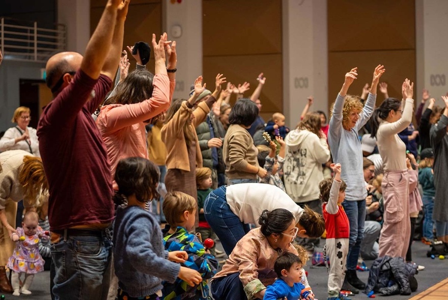An audience of small childrent and their carers with hands in the air. 