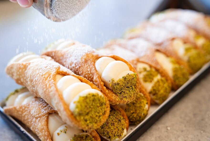 Pistachio and ricotta cannolis on a tray, dusted with icing sugar.