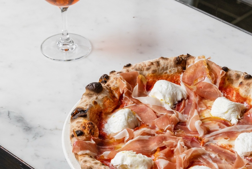 A traditional-style Italian pizza with buffalo mozzarella and prosciutto, a glass of rose wine in the background.