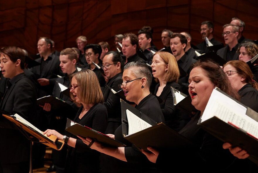 Massed choir dressed in black holding open musical scores as they sing.