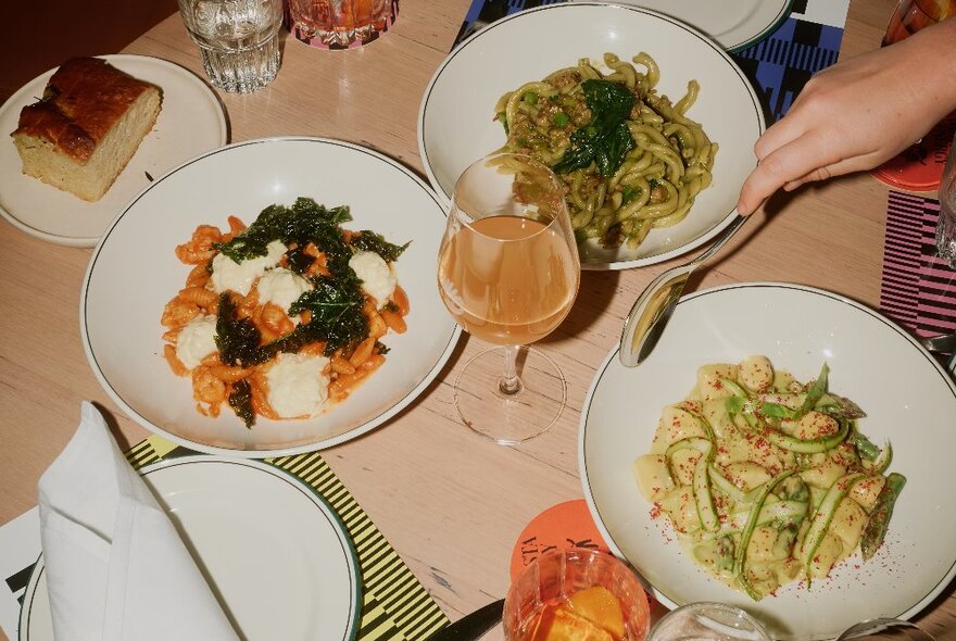 Three bowls of pasta, plus glasses of wine, cutlery and napkins on a wooden dining table in a restaurant, with a hand reaching in with a spoon.