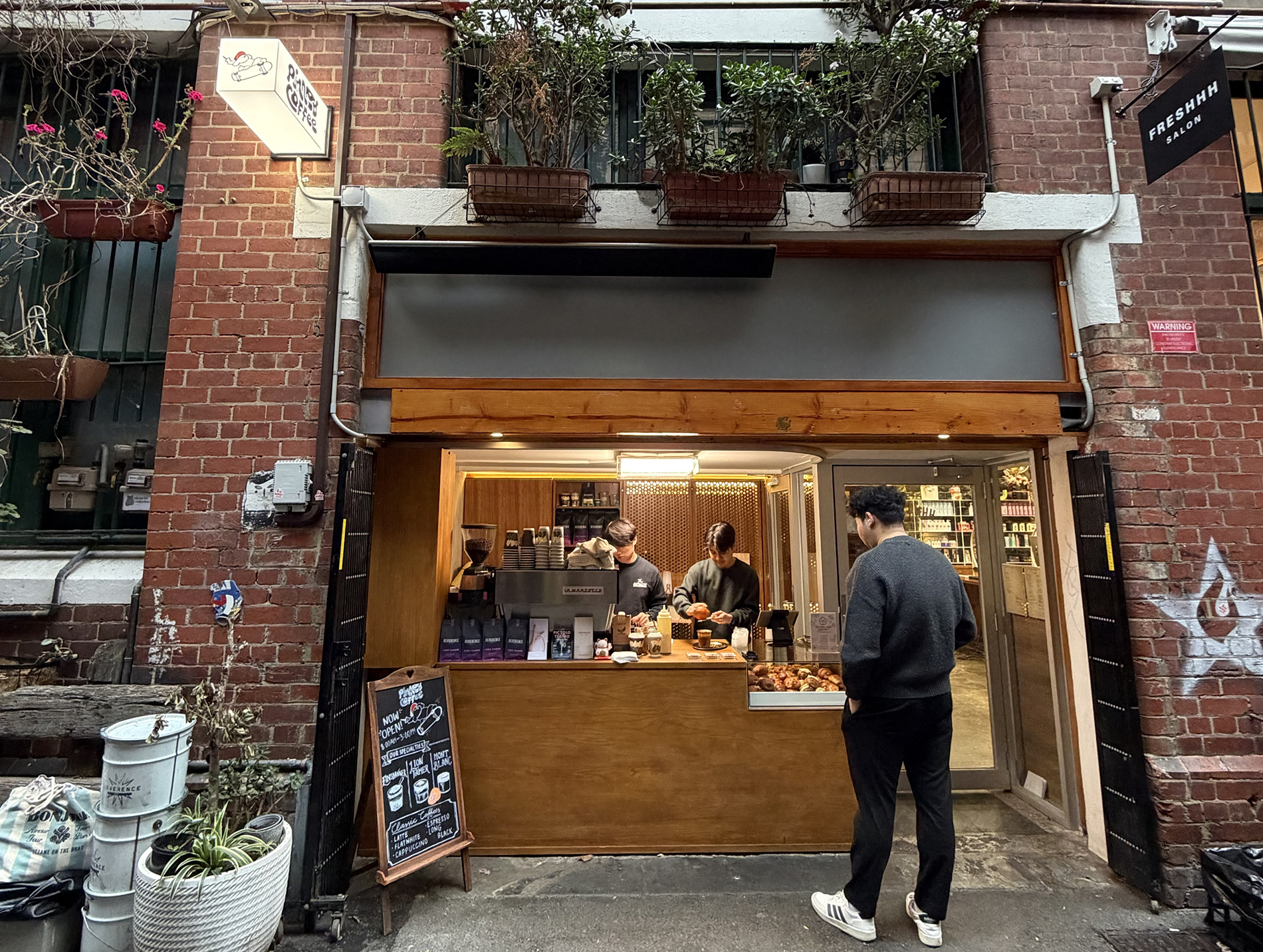 A man ordering at a coffee window with a blackboard menu and plants.