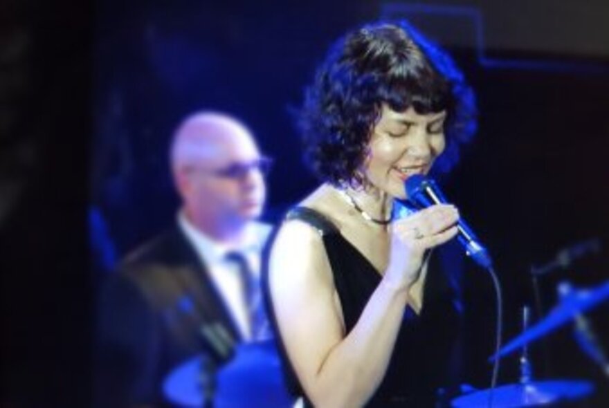 Singer Jackie Bornstein singing live holding a microphone and with a blurred musician behind her.
