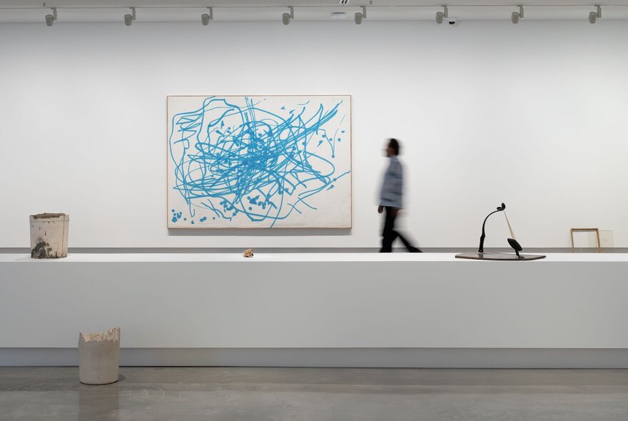 A large white room in a gallery, with a person in the background walking past a large contemporary artwork hanging on the wall.
