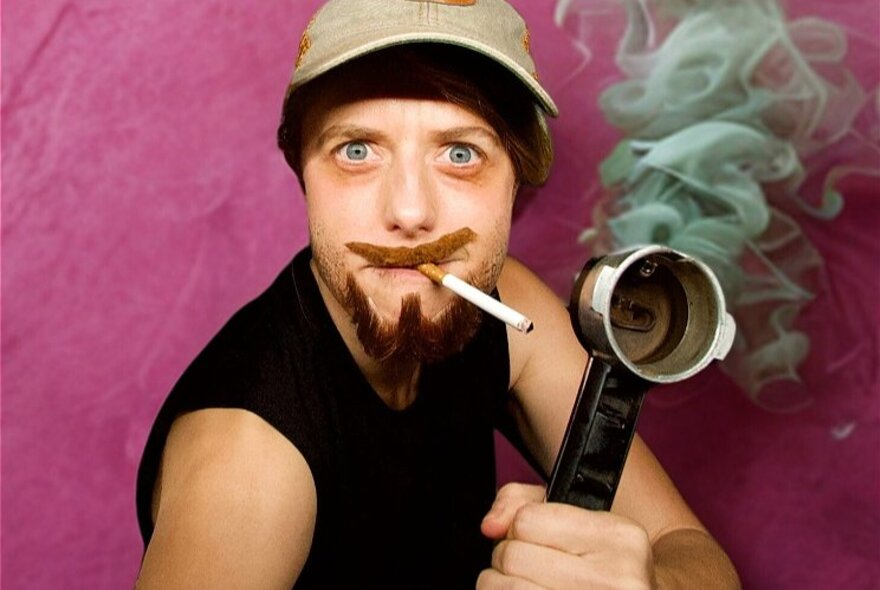 Performer in drag with a fake mustache and beard, smoking a cigarette and holding a coffee making head from a coffee machine.