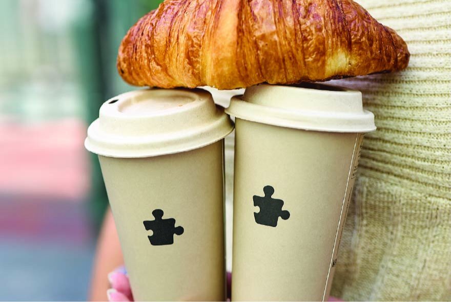 Two takeaway coffee cups and a croissant being held by a hand and balanced against a person's chest. 