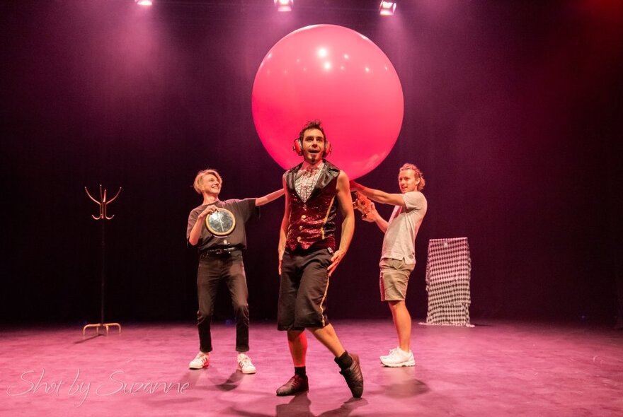 The Quizzical Mr Jeff on stage performing with a large red balloon behind him and two child volunteers.