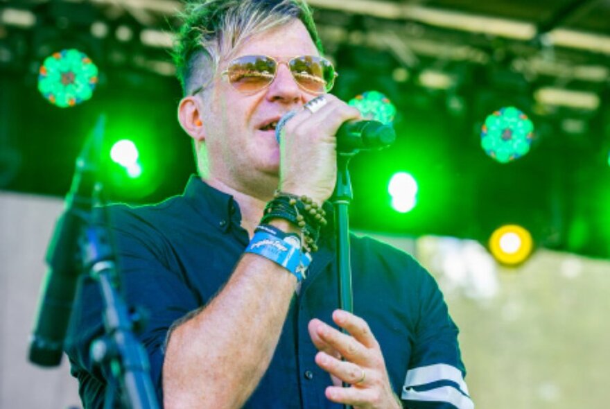 A singer wearing aviator sunglasses performing on stage with green lights behind him. 