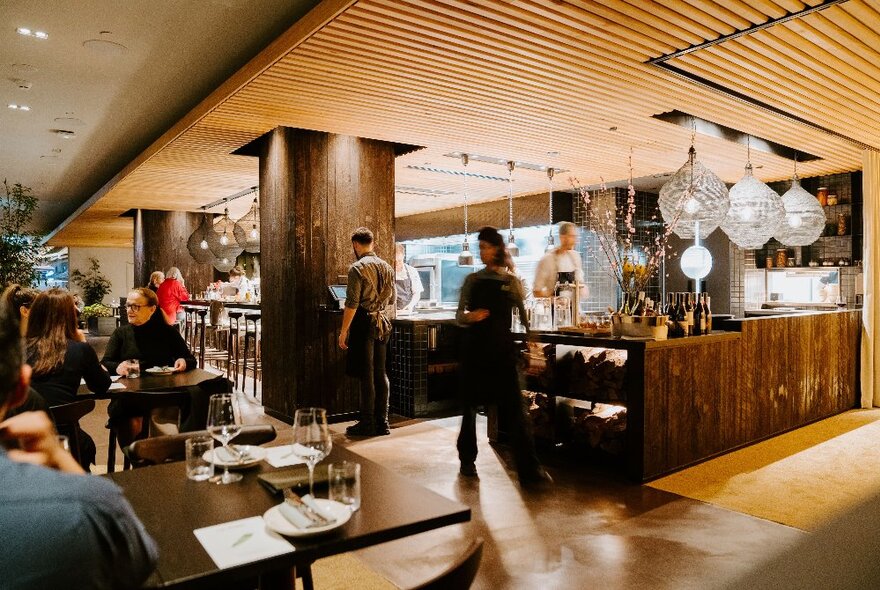 Modern wood-toned interior of a restaurant with staff in the background and dining patrons seated at tables.