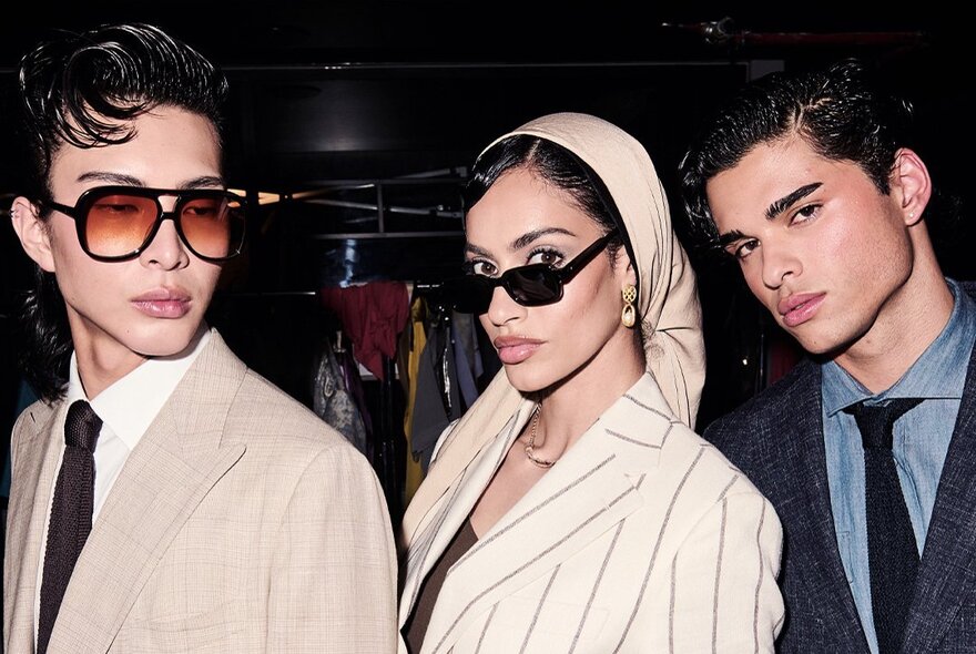 Two fashion models posing backstage in tailored neutral-toned blazers and chic dark sunglasses, with a third fashion model wearing a dark suit.
