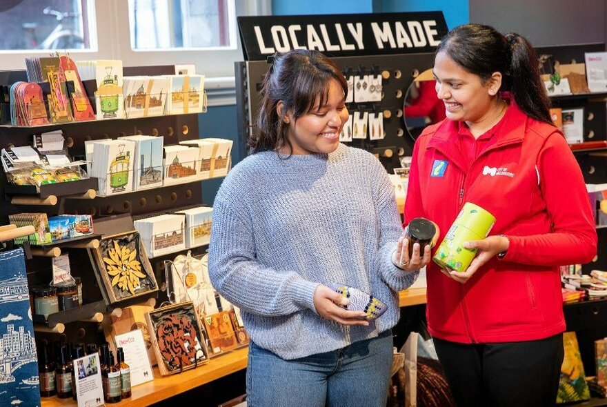 Two people in a local handicrafts store, one in a red uniform showing the other some products.
