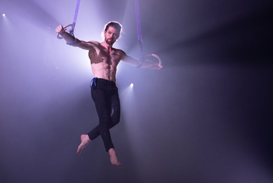 A bare-chested man in dark leggings, using rings and ropes to hold his body aloft in mid air, spotlit from behind.