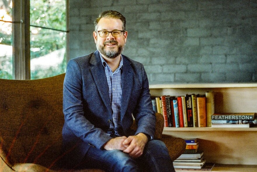 A smiling man seated in a room with a low bookshelf and grey brick wall. 