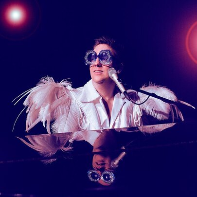 A person dressed as Elton John, with big glasses and feathered shoulder pads, seated at a piano on stage with two spotlights. 