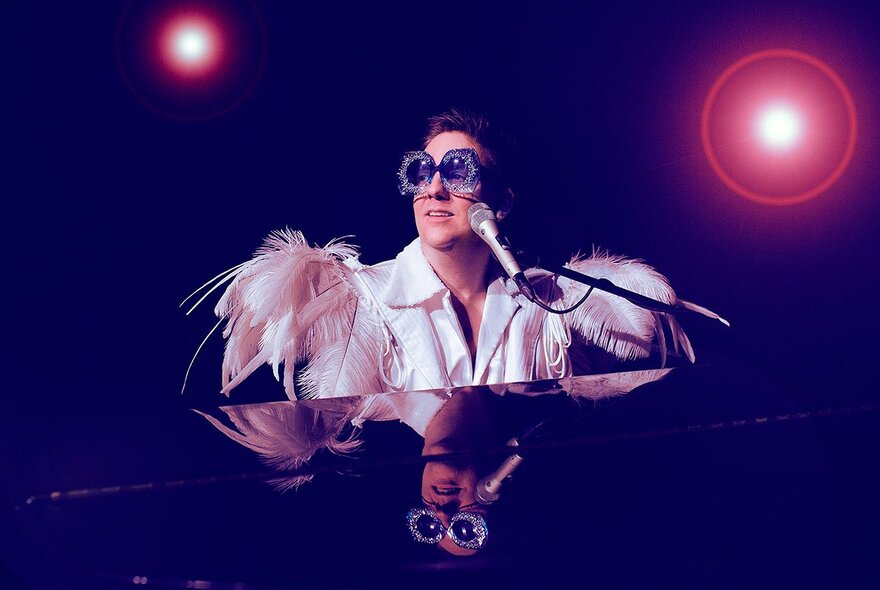 A person dressed as Elton John, with big glasses and feathered shoulder pads, seated at a piano on stage with two spotlights. 