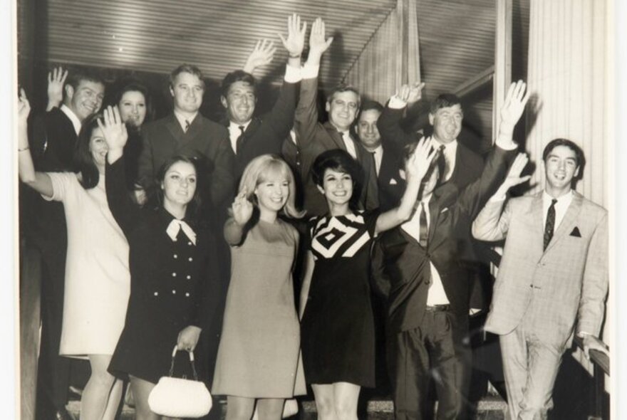 A large group of people, dressed in 1960s fashion, waving enthusiastically at the camera; black and white image.
