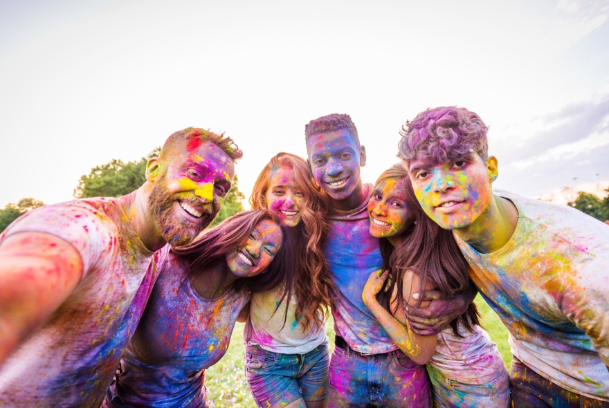 A group of six friends smiling outdoors, covered in coloured powder at a Holi Festival and posing for a selfie. 