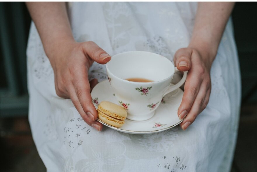 Detail of hands holding a cup of tea and macaron, wearing a white gown.