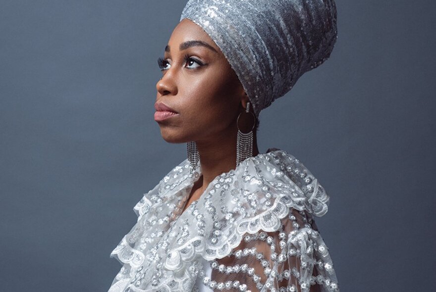 Singer Jazzmeia Horn standing in profile, her head slightly tilted and looking up, wearing a silver turban and a delicately ruffled and textured white lace top.