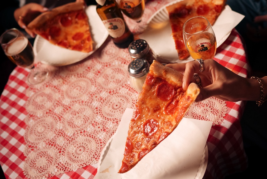 A woman's hand holding a slice of New York-style pepperoni pizza over a table with a red and white checked tablecloth.