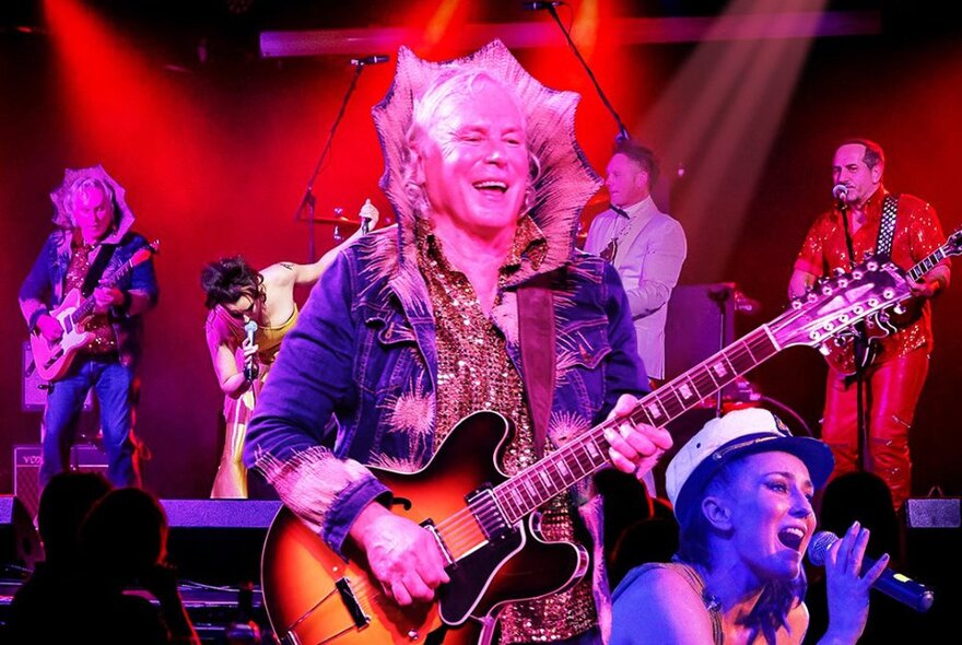 A band in dressed in glam-rock costumes performing on a red-lit stage.