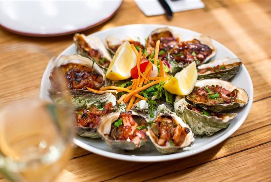 Looking through a blurred wineglass on the left to a plate of oysters Kilpatrick with wedges of lemon.
