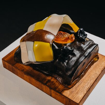 Small sculpture made from coloured glass displayed on a wooden base and white pedestal.