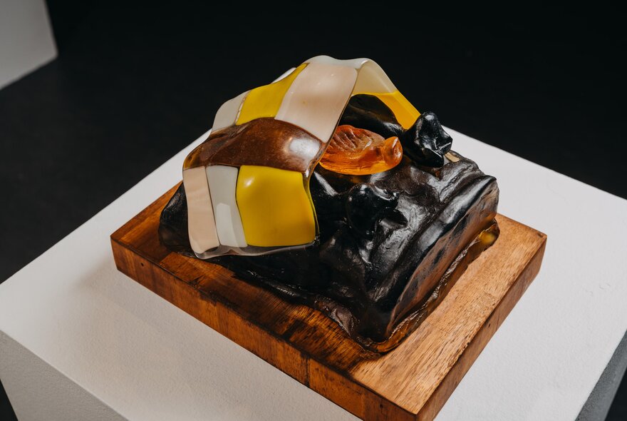 Small sculpture made from coloured glass displayed on a wooden base and white pedestal.