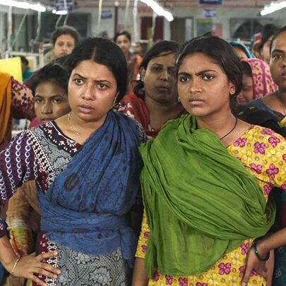 A factory room full of Bangladeshi women in traditional clothing all looking straight ahead, annoyed. 