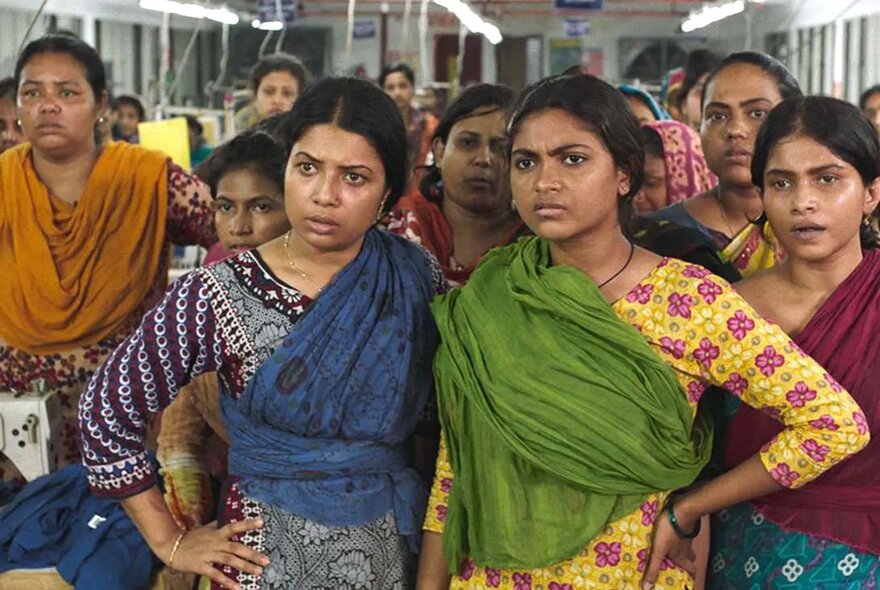 A factory room full of Bangladeshi women in traditional clothing all looking straight ahead, annoyed. 
