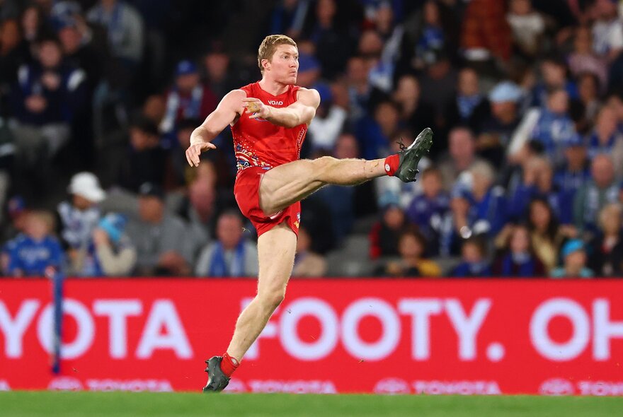 A Gold Coast SUNS AFL player with one leg raised high, having just kicked a football, a blurred stadium crowd in the background behind him. 
