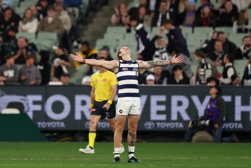 A Geelong AFL player celebrating on the field after having scored, his arms outstretched, with blurred fans in the stadium in the background and an umpire walking behind him.