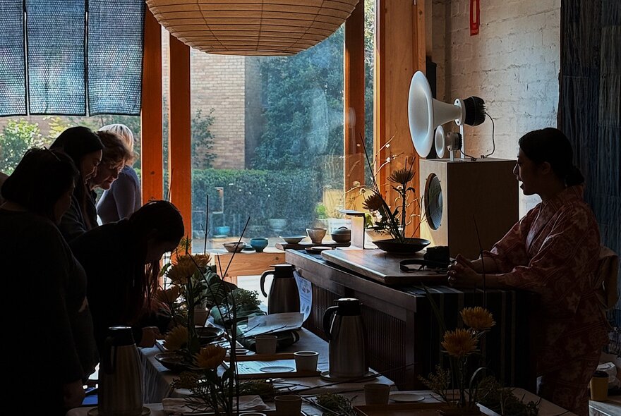 A group of people participating in a traditional Japanese flower arranging Ikebana workshop in a dimly lit gallery or studio space.