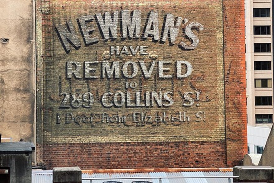 A ghost sign on a brick wall, faded and from and earlier time, the painted words NEWMAN'S HAVE REMOVED TO 289 COLLINS ST still just visible. 