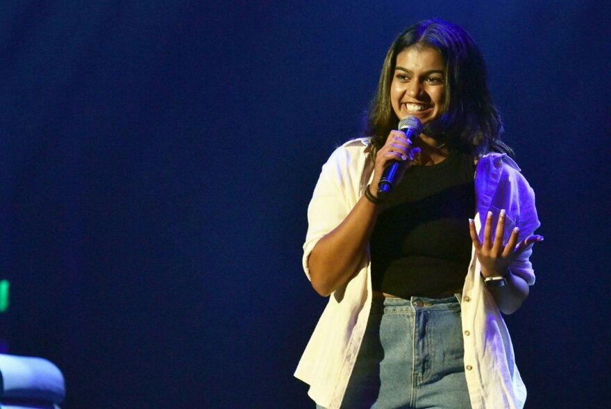 A teenager smiling and holding a microphone in her hand, performing stand up comedy on a stage.