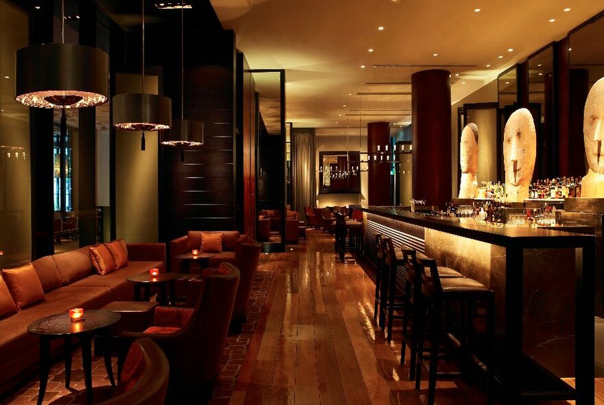 Interior of Ru-Co Bar showing high bar stools underneath the long dark bar counter, and smaller intimate tables and chairs against the wall, wooden floors and soft lighting. 