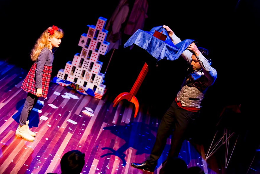 A magician on a stage, holding a blue scarf over an object, interacting with a primary school aged child who also stands on the stage.