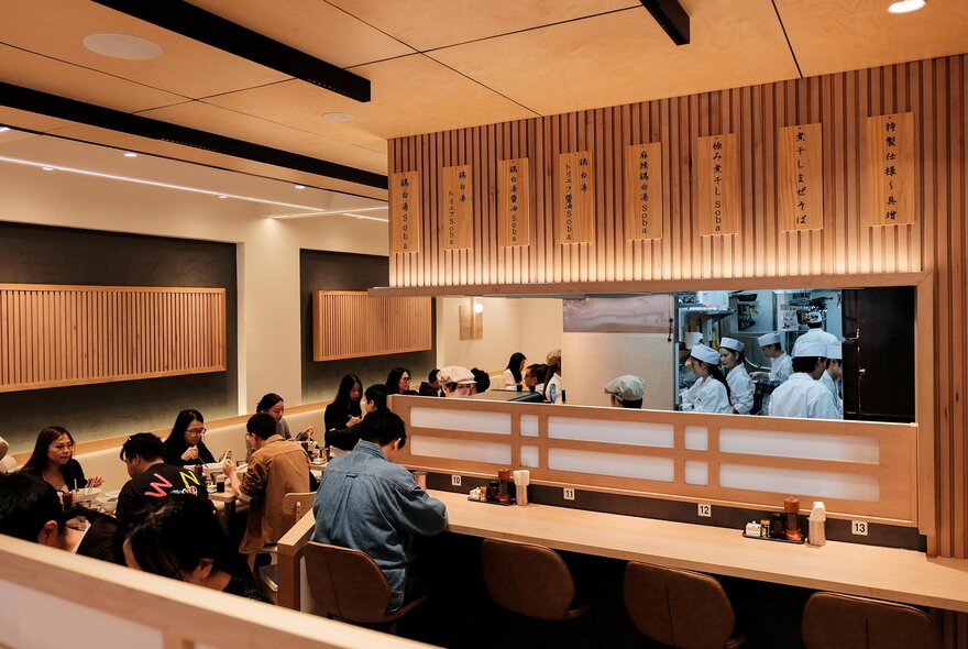 The warm timber interior of Ginza Kagari restaurant in Melbourne, with patrons eating at tables and along a narrow bar facing the kitchen.