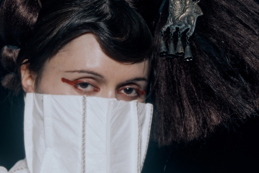 A person with a distinctive makeup style, dark hair arranged in an elaborate updo, with the lower part of their face covered by a white, structured garment.