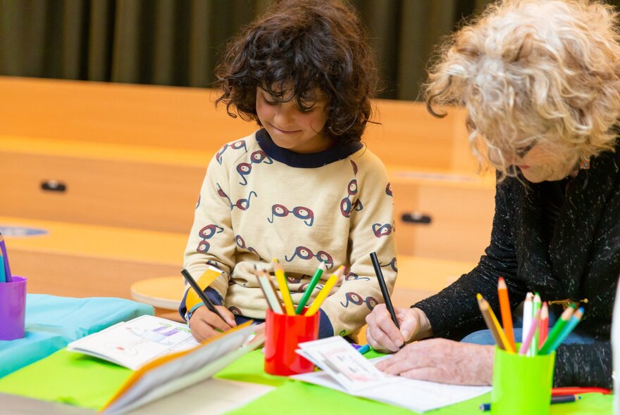 A young child working at a colourful desk with an older adult working next to him.
