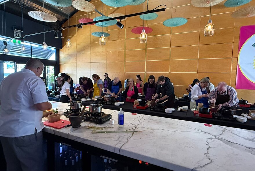People lined up at workstations with Asian cooking equipment in front of a chef at a marble counter, in a restaurant with ceiling hung with paper umbrellas.