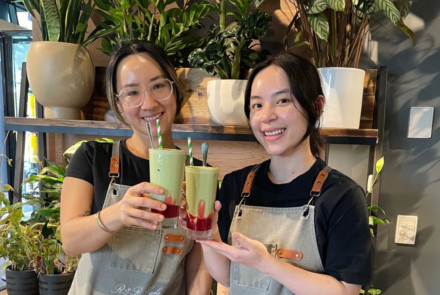 Two smiling cafe staff holding green drinks in front of a wall of green plants inside a cafe.