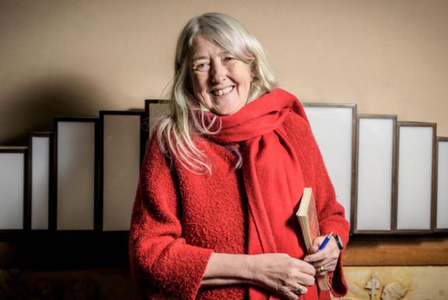 Portrait of author and classicist Mary Beard, smiling as she holds a book and a pen.